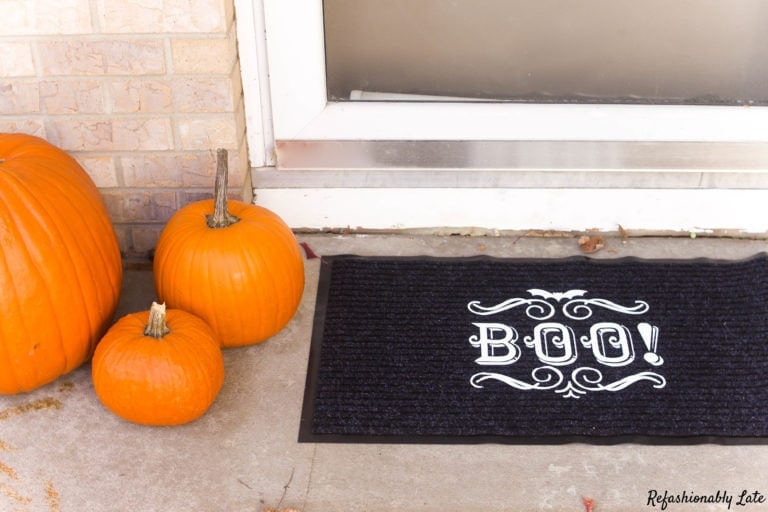 black doormat with the word BOO written on top sitting by 3 pumpkins on the front porch