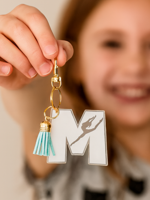 Personalized gymnast “M” acrylic keychain held by a smiling child, featuring a clear engraved design with a gymnast silhouette, gold hardware, and a soft teal tassel—perfect for gym bags, backpacks, and showing off team spirit.