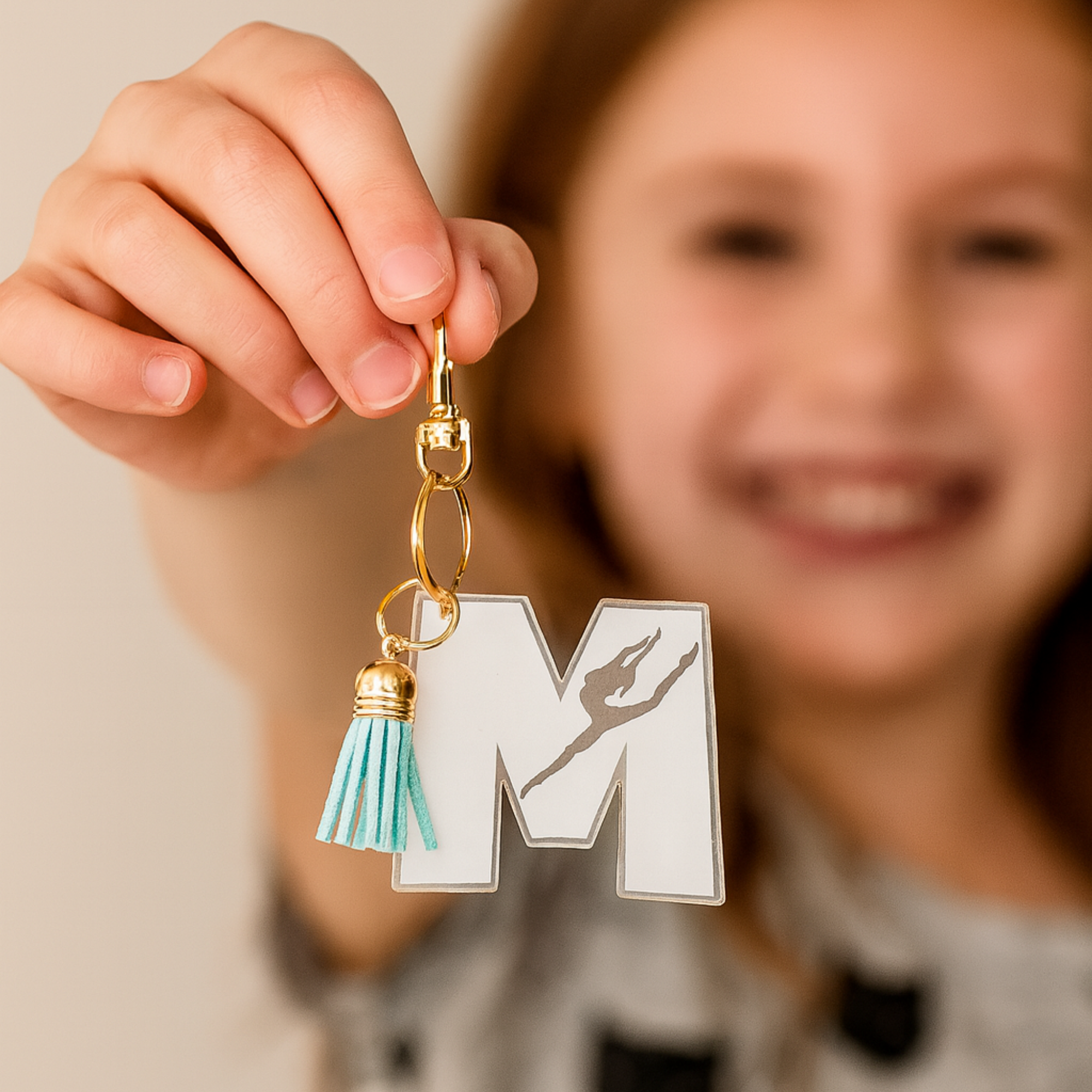 Personalized gymnast “M” acrylic keychain held by a smiling child, featuring a clear engraved design with a gymnast silhouette, gold hardware, and a soft teal tassel—perfect for gym bags, backpacks, and showing off team spirit.
