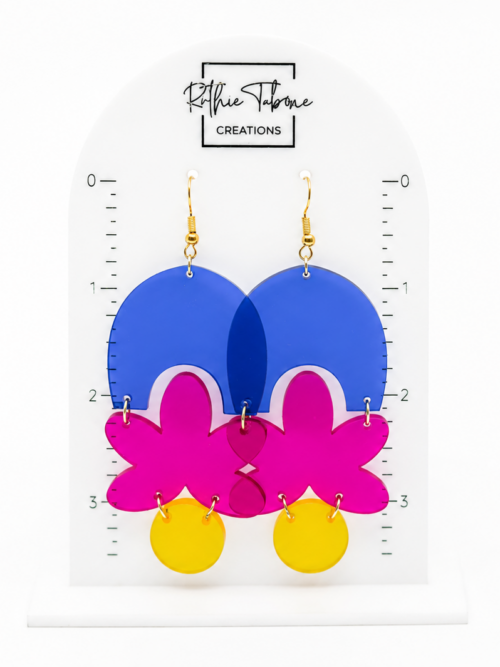 Colorful handmade acrylic dangle earrings featuring blue arches, bright pink floral shapes, yellow circles, and gold hooks displayed on an earring size guide.