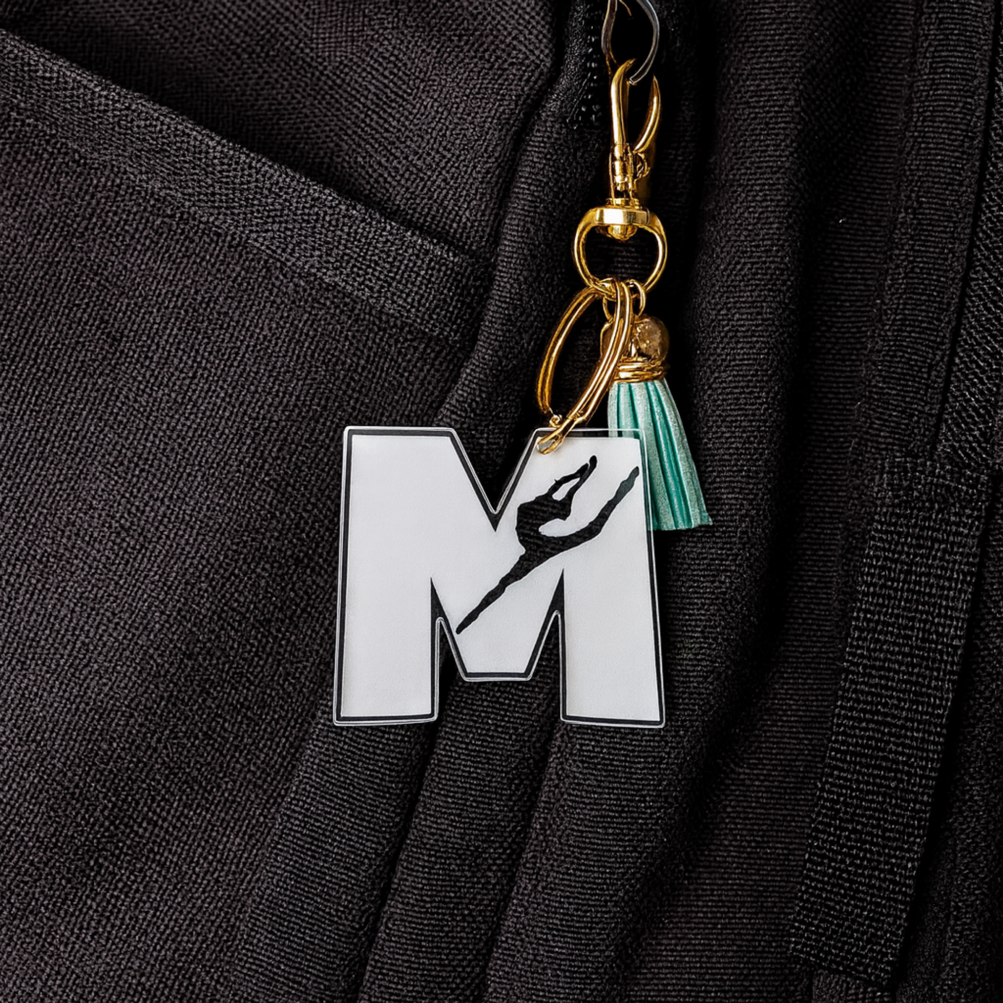 Personalized gymnast “M” acrylic keychain clipped onto a backpack, featuring a clear engraved letter with a gymnast silhouette, gold hardware, and a teal tassel—perfect for adding a custom, sporty touch to gym bags and everyday gear.