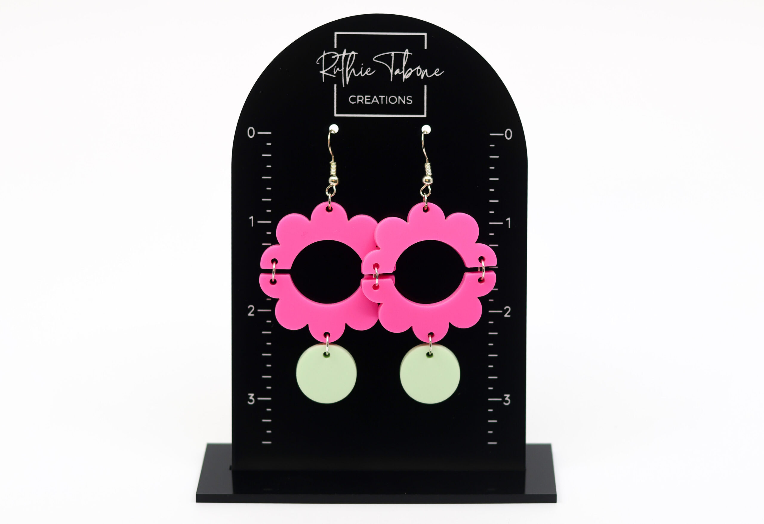 Hot pink flower dangle earrings with mint green accents displayed on a black measurement stand, highlighting their bold contrast, playful shape, and approximate drop length for easy size reference.