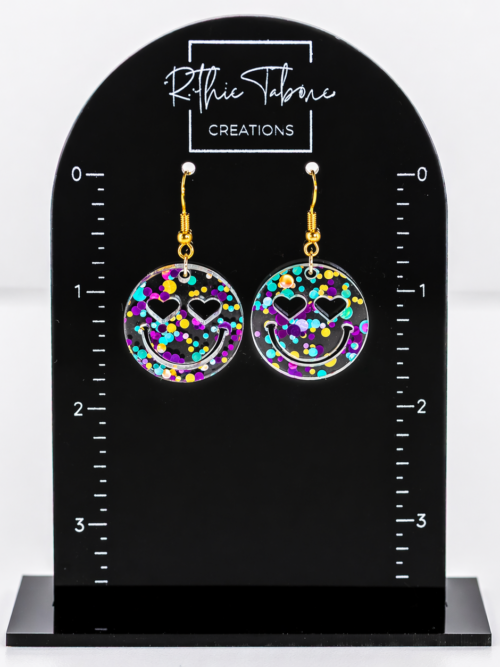 Clear acrylic smiley face dangle earrings with colorful confetti and heart-shaped eyes, finished with gold hooks, displayed on a black Ruthie Tabone Creations earring card with measurement markings.
