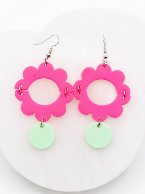 Vibrant hot pink acrylic flower dangle earrings with a bold open-circle center and soft mint green drop accents, styled on a white stone backdrop for a fun, eye-catching, and modern statement look.