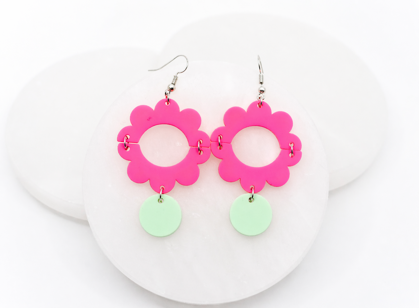 Vibrant hot pink acrylic flower dangle earrings with a bold open-circle center and soft mint green drop accents, styled on a white stone backdrop for a fun, eye-catching, and modern statement look.