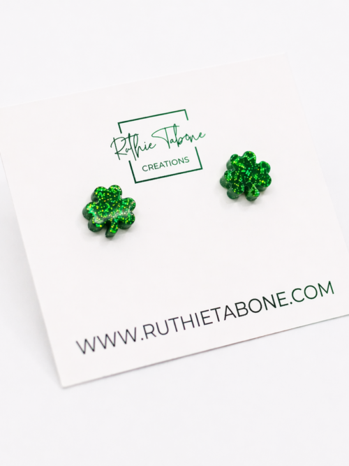 Green glitter shamrock stud earrings on a white Ruthie Tabone Creations display card, photographed on a bright white background with a clean, minimalist style.