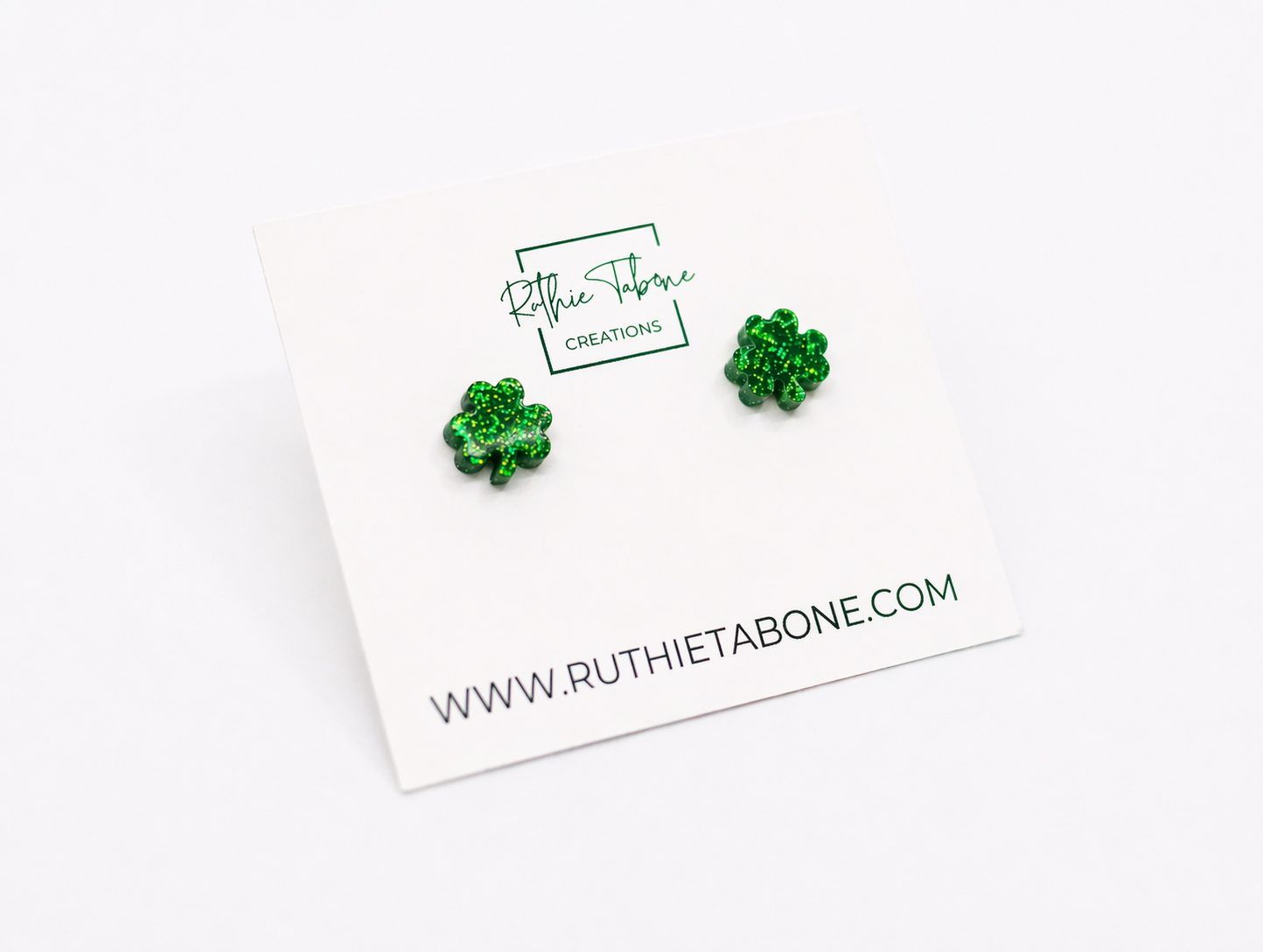 Green glitter shamrock stud earrings on a white Ruthie Tabone Creations display card, photographed on a bright white background with a clean, minimalist style.