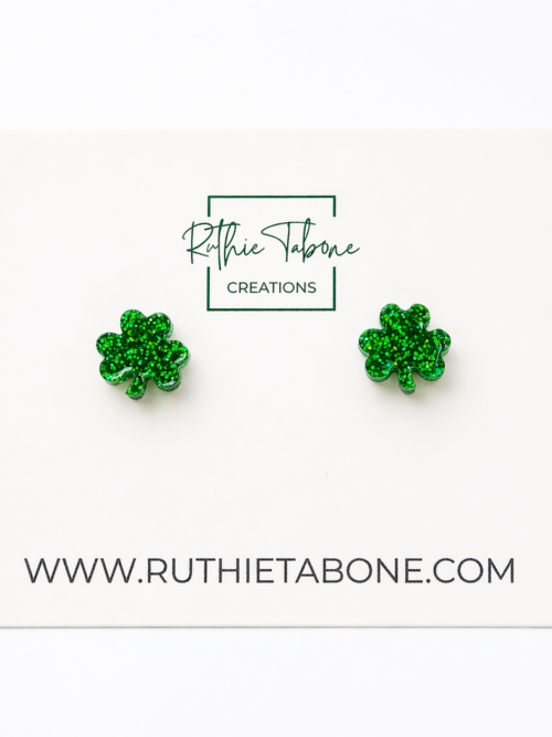 Bright, high-key product photo of green glitter shamrock stud earrings displayed on a white Ruthie Tabone Creations card, with a clean white background and the website URL printed below.