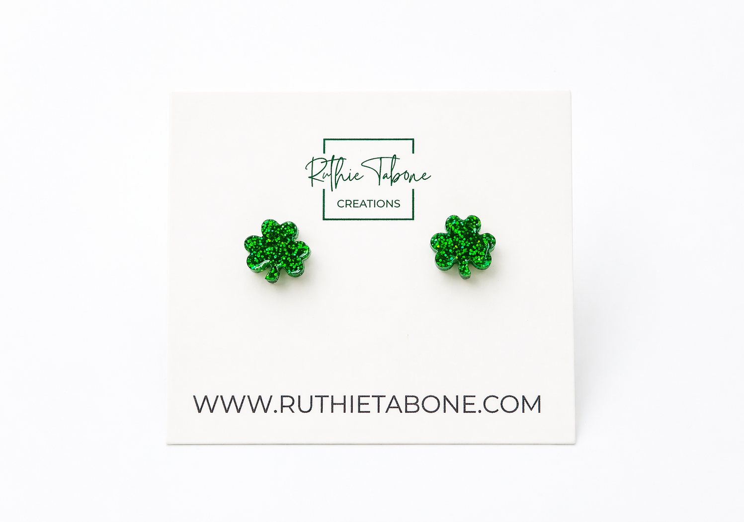 Bright, high-key product photo of green glitter shamrock stud earrings displayed on a white Ruthie Tabone Creations card, with a clean white background and the website URL printed below.