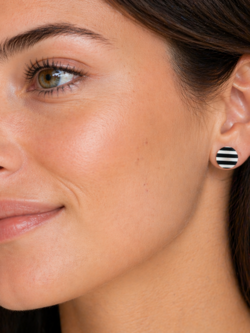 Close-up of a woman wearing black and white striped round stud earrings, shown in side profile with natural makeup and soft lighting against a neutral background.