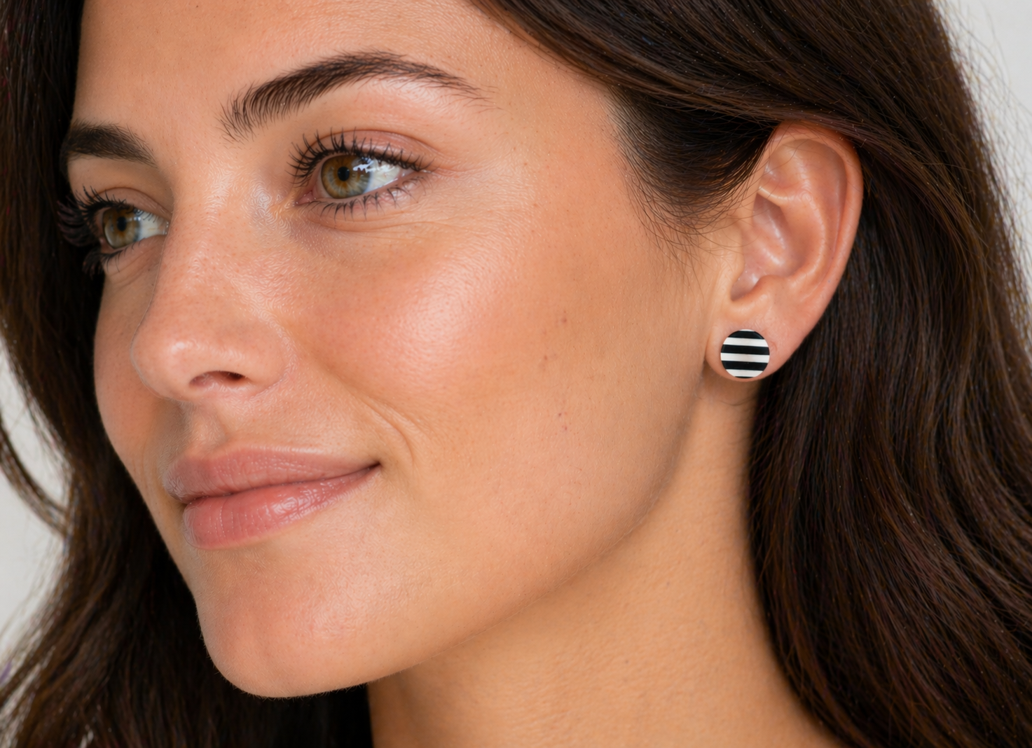 Close-up of a woman wearing black and white striped round stud earrings, shown in side profile with natural makeup and soft lighting against a neutral background.
