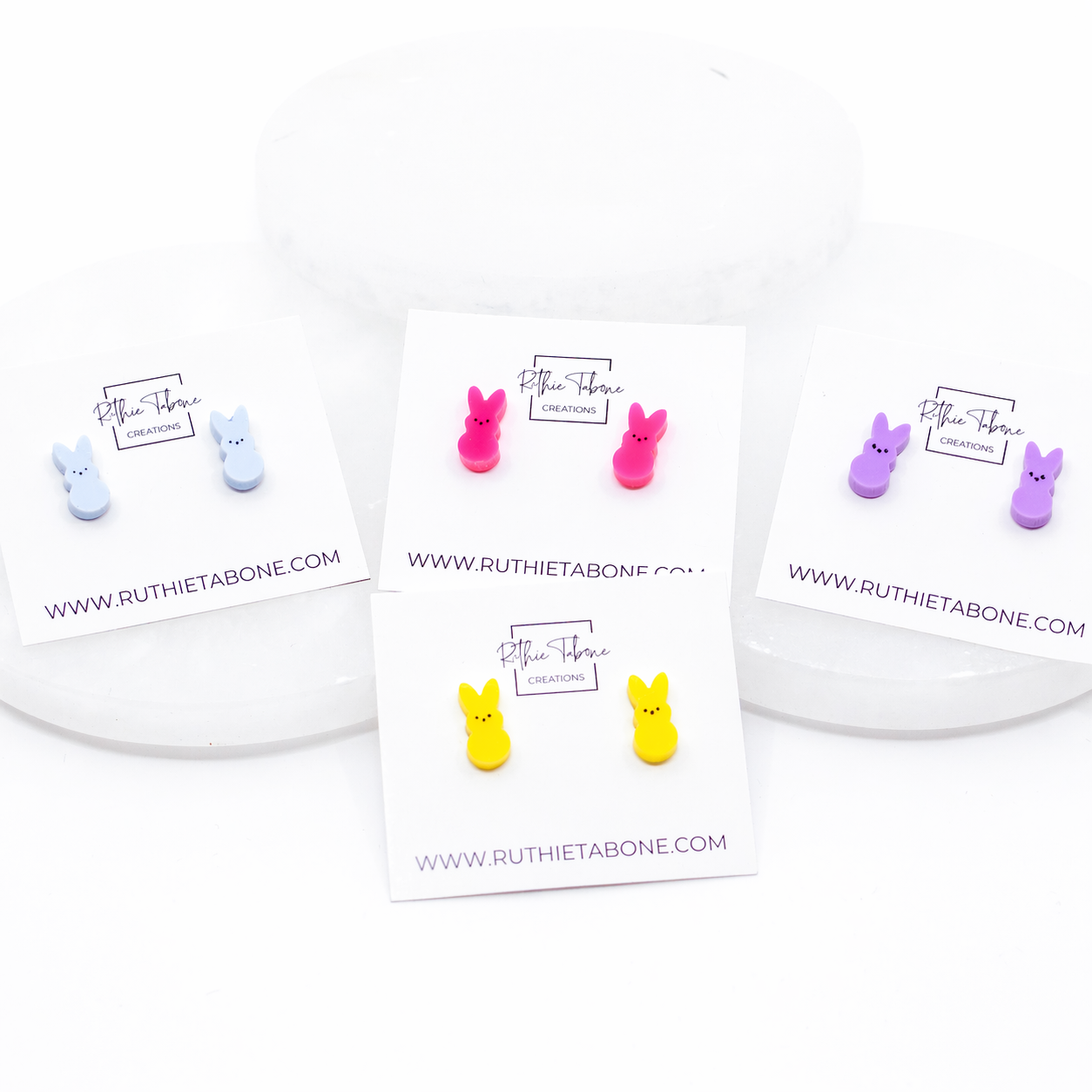 Pastel bunny stud earrings in yellow, pink, lavender, and light blue displayed on Ruthie Tabone Creations cards in a styled, angled product layout with white risers.