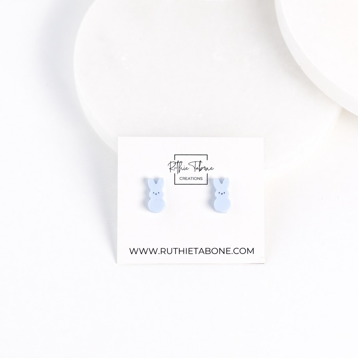 Light blue bunny stud earrings displayed on a branded Ruthie Tabone Creations card, styled on a clean white background with soft, minimal props.