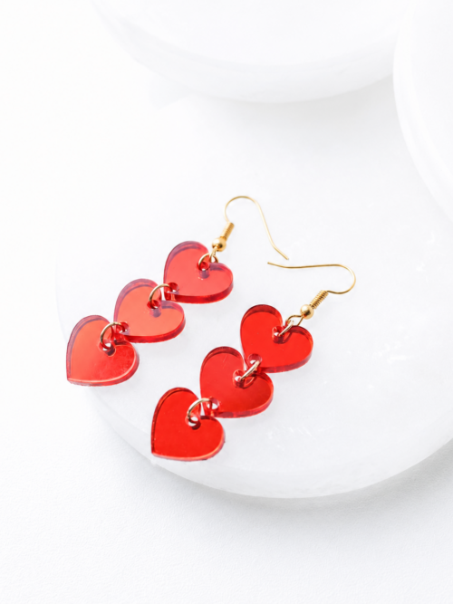 image alt text Red acrylic heart dangle earrings featuring three linked translucent hearts with gold-tone jump rings and hypoallergenic gold hooks, displayed on a bright white background.