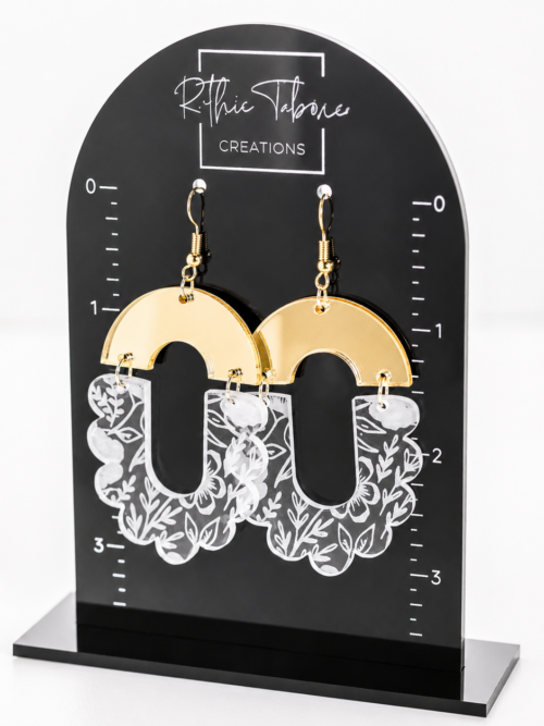 Gold mirror acrylic and clear floral engraved statement earrings displayed on a black Ruthie Tabone Creations earring stand. These lightweight acrylic dangle earrings feature a modern arch design with white floral engraving, gold hypoallergenic hooks, and a bold boho-inspired style.