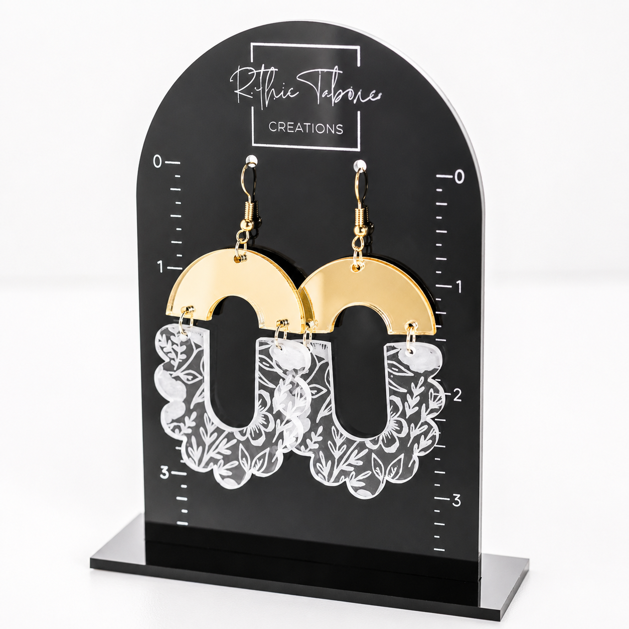 Gold mirror acrylic and clear floral engraved statement earrings displayed on a black Ruthie Tabone Creations earring stand. These lightweight acrylic dangle earrings feature a modern arch design with white floral engraving, gold hypoallergenic hooks, and a bold boho-inspired style.