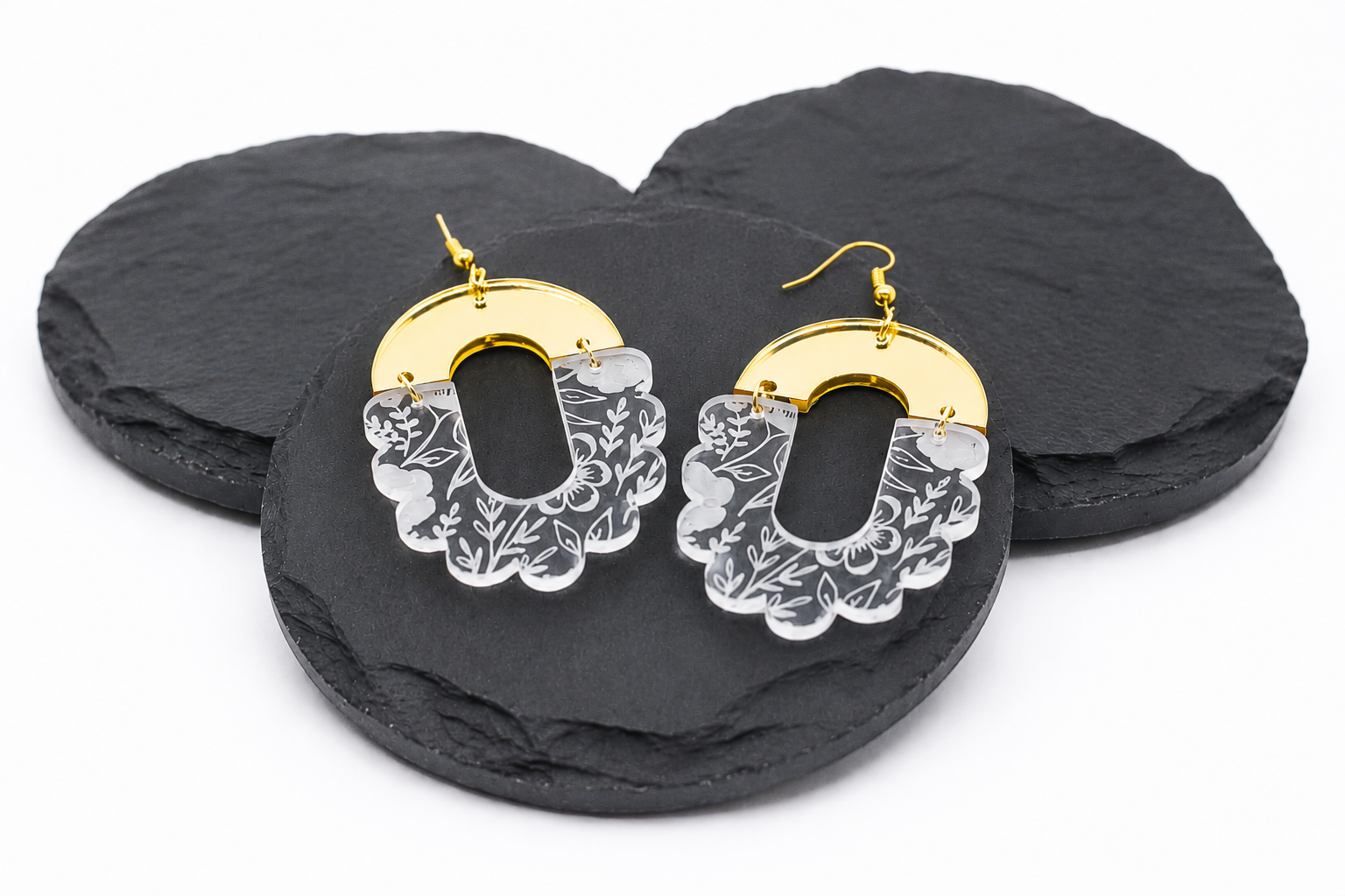 Gold mirror acrylic and clear floral engraved statement earrings displayed on black slate coasters. These lightweight acrylic dangle earrings feature a modern arch shape with scalloped floral detailing, gold hypoallergenic hooks, and an elegant boho-inspired design.