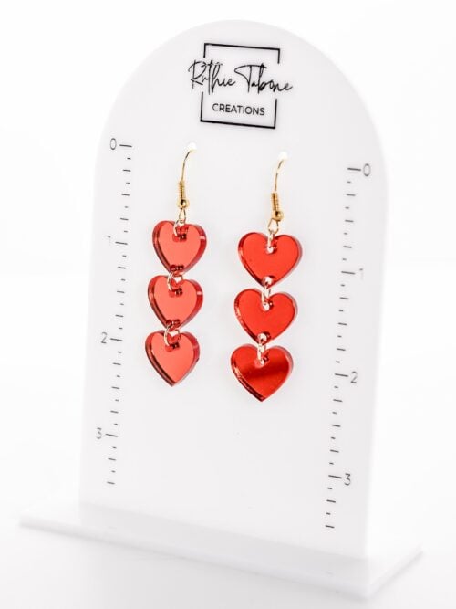 Red acrylic heart dangle earrings featuring three linked translucent hearts with gold-tone hypoallergenic hooks, displayed on a Ruthie Tabone Creations earring stand with size measurements.