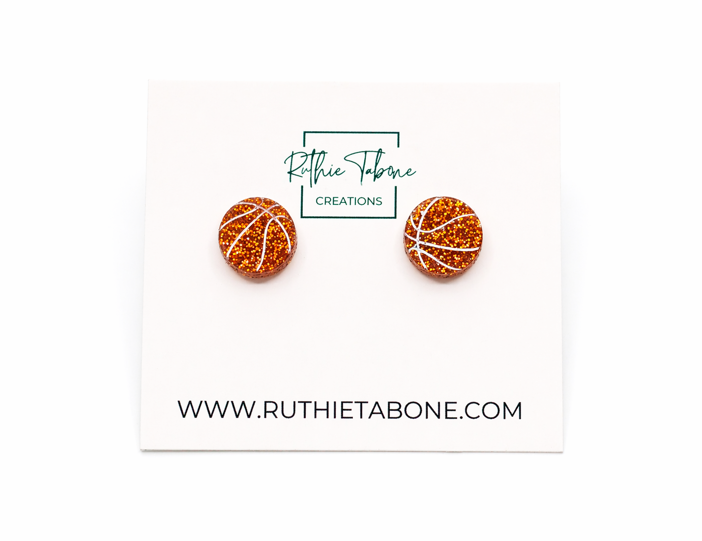 Glitter basketball stud earrings with white line detailing displayed on a Ruthie Tabone Creations card, photographed on a clean white background.