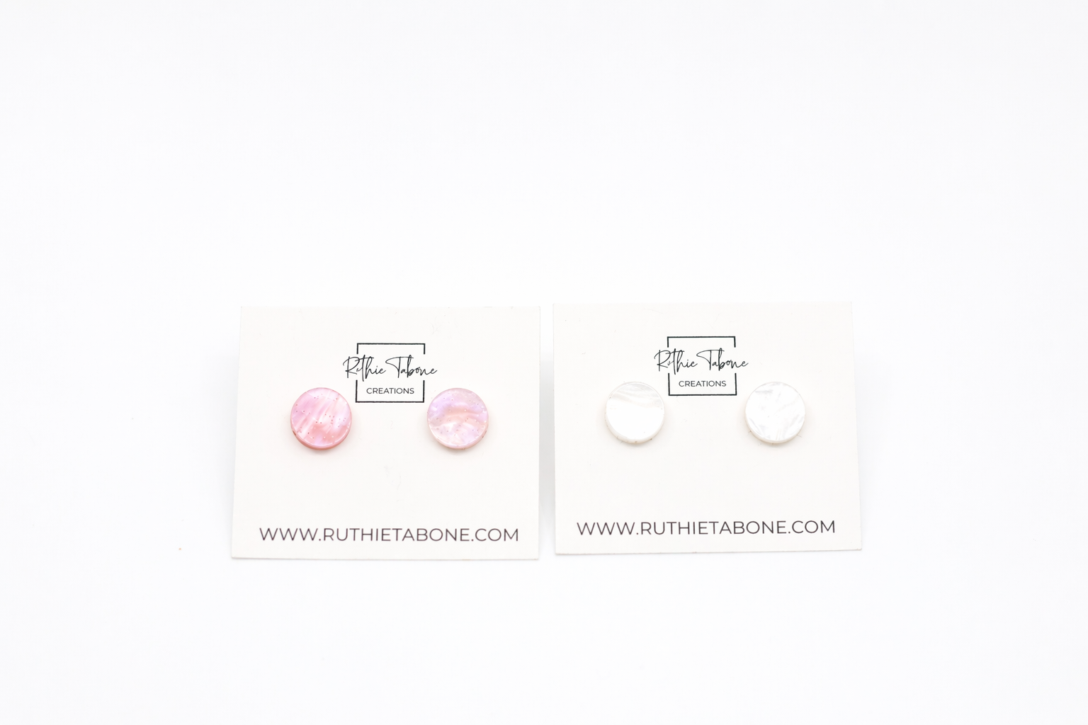 Two pairs of round stud earrings—one in pink marbled acrylic and one in white marbled acrylic—displayed on Ruthie Tabone Creations cards against a clean white background.