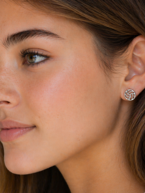 Close-up side profile of a woman wearing silver volleyball stud earrings, with natural makeup and soft lighting highlighting the jewelry against a neutral background.