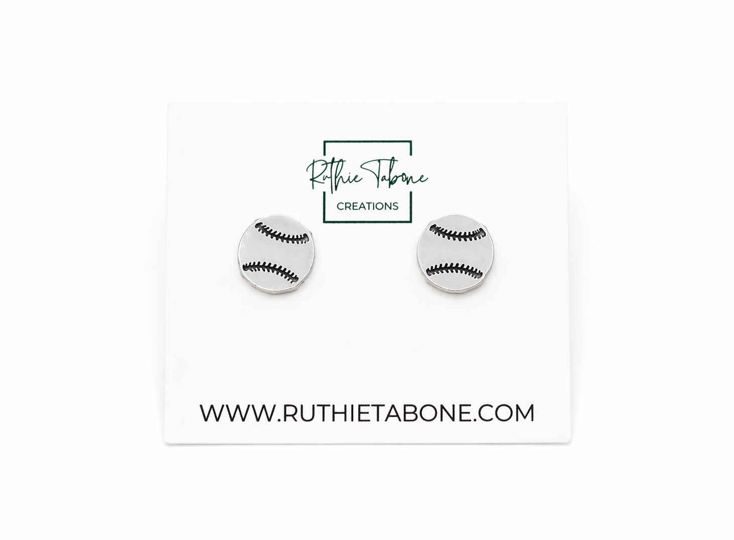 Silver baseball stud earrings displayed on a branded Ruthie Tabone Creations card, featuring a round design with engraved black stitching details for a sporty, minimalist look.