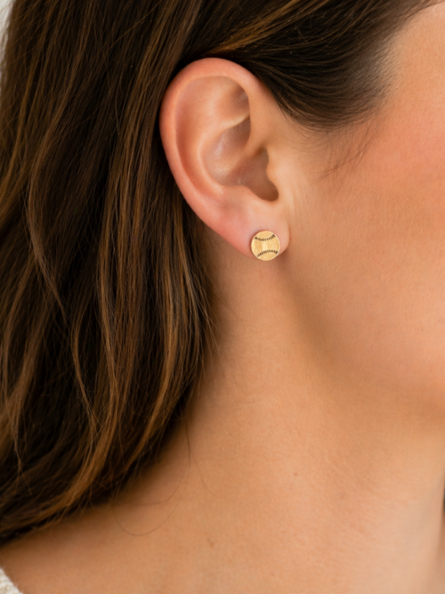 Close-up of a woman wearing gold baseball stud earrings, featuring a round design with engraved stitching detail on her ear.