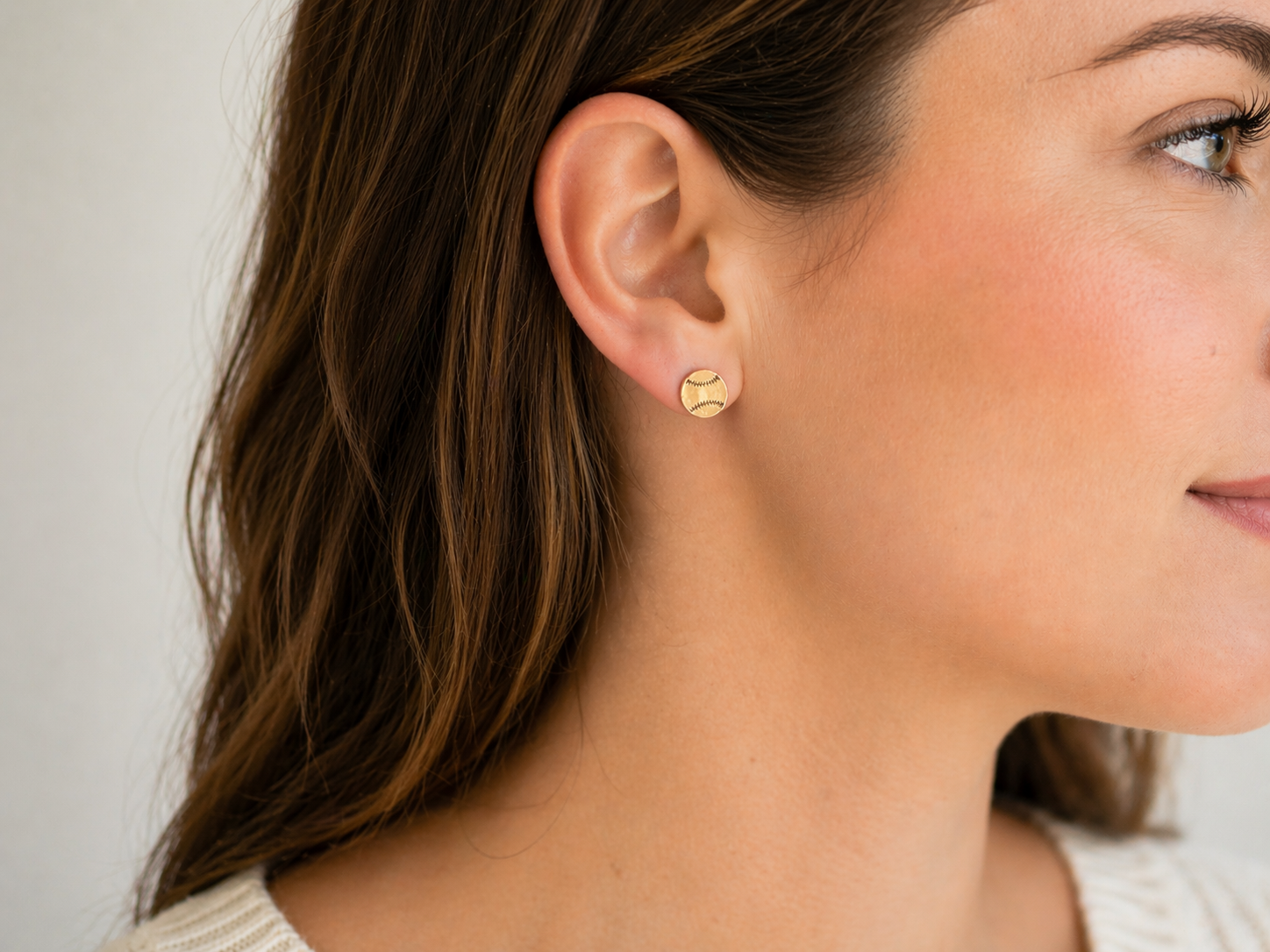 Close-up of a woman wearing gold baseball stud earrings, featuring a round design with engraved stitching detail on her ear.