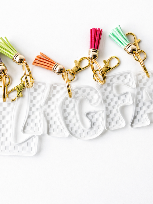 Clear acrylic checkerboard initial keychains spelling “LKGFA,” finished with gold hardware and bright tassel accents—fun, customizable accessories perfect for backpacks, keys, or gifts.