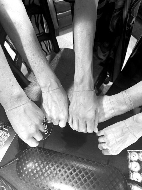 Black-and-white close-up of five wrists wearing delicate permanent bracelets, gathered together over a tabletop in a celebratory pose. The minimalist bracelets symbolize friendship and lasting connections, while a jeweler's work mat and tools in the background hint at the permanent jewelry experience.