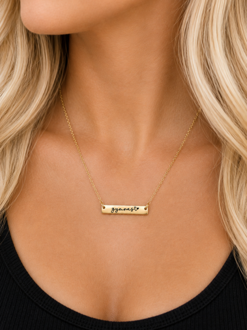 Gold gymnast bar necklace with black script “gymnast” and heart engraving, modeled on a delicate gold chain.